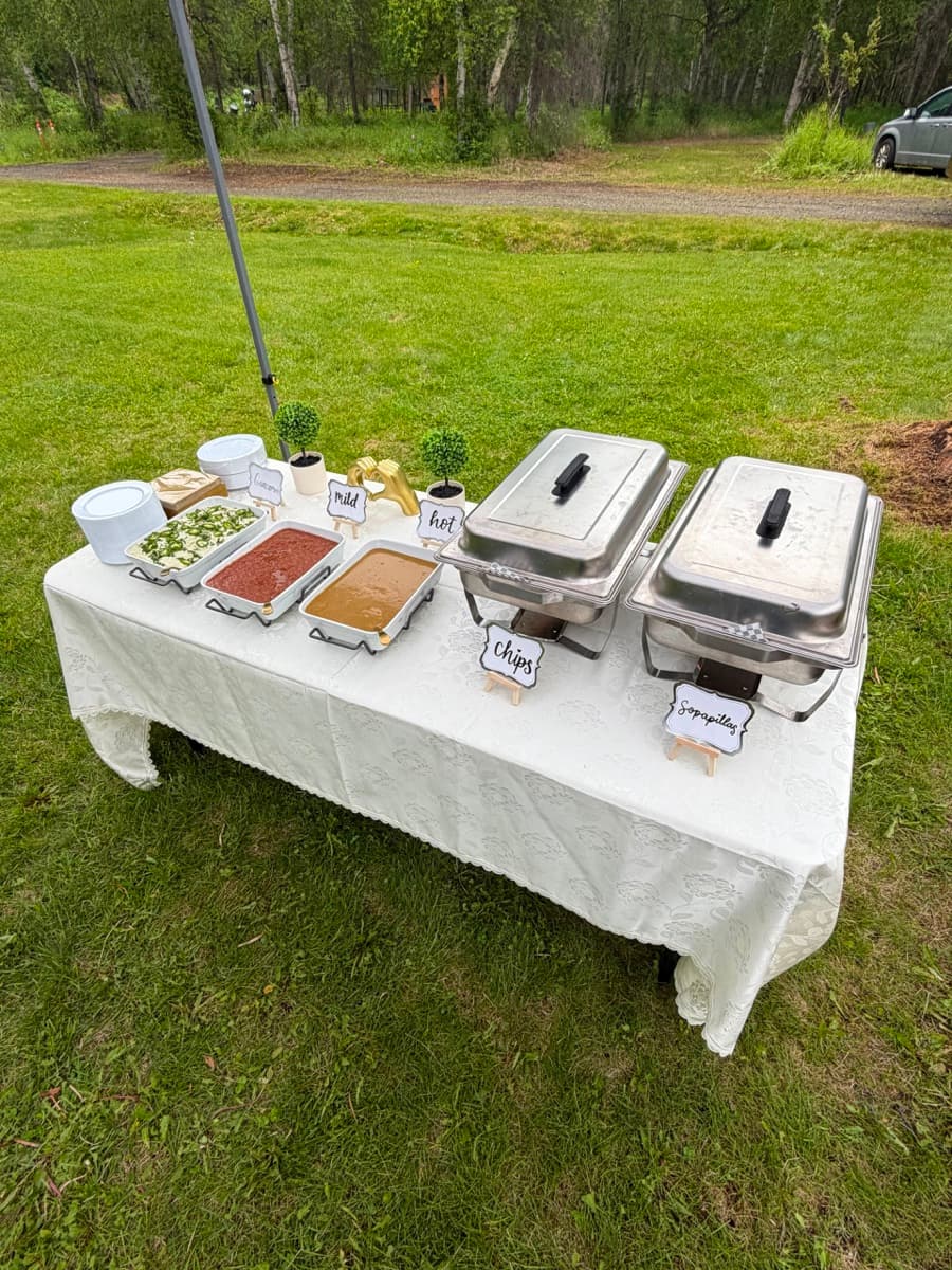 Complete outdoor buffet table setup with lace tablecloth, warmers and fresh salsas